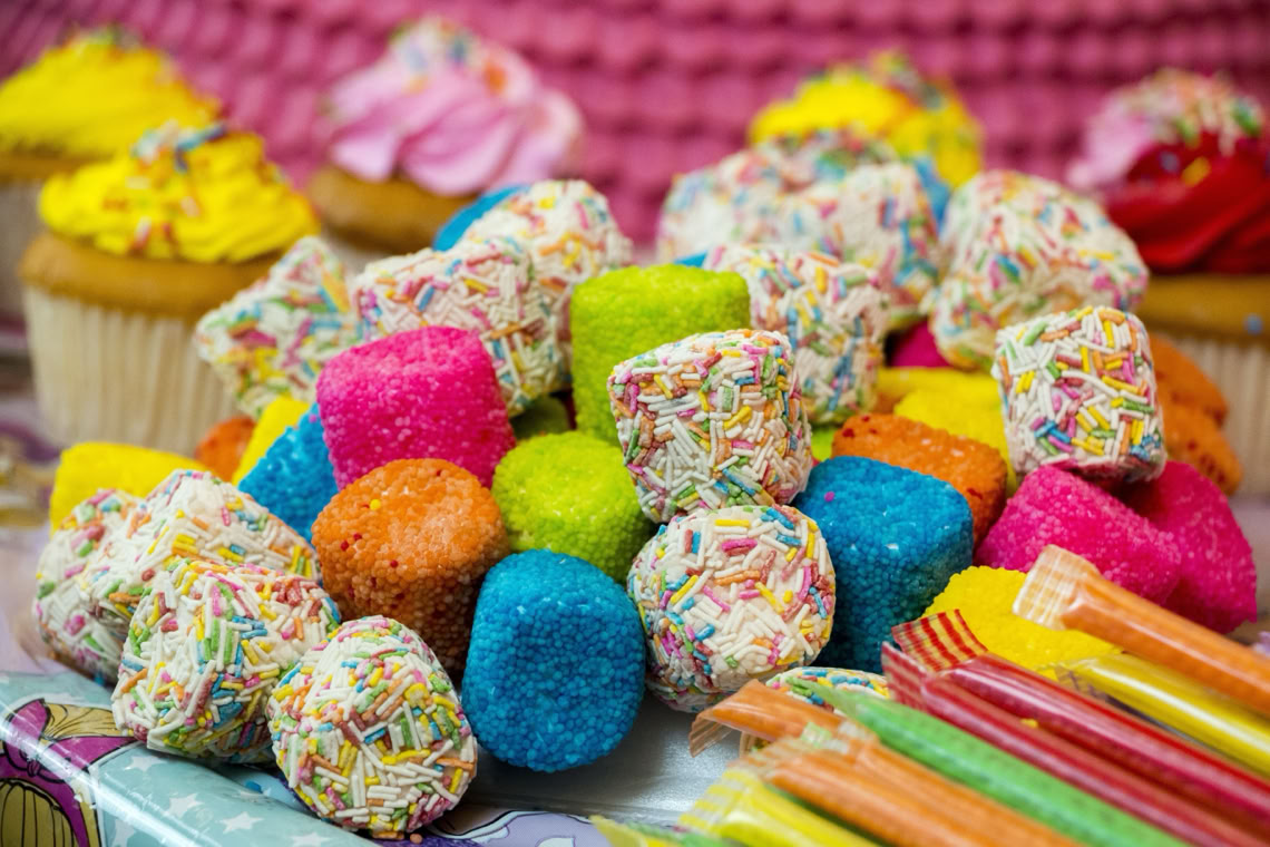 Close-up van een kleurrijke selectie van Chris Candy Corner's snoepgoed, inclusief zachte marshmallows met discodip en felgekleurde suikersnoepjes, met op de achtergrond cupcakes en drinkrietjes.